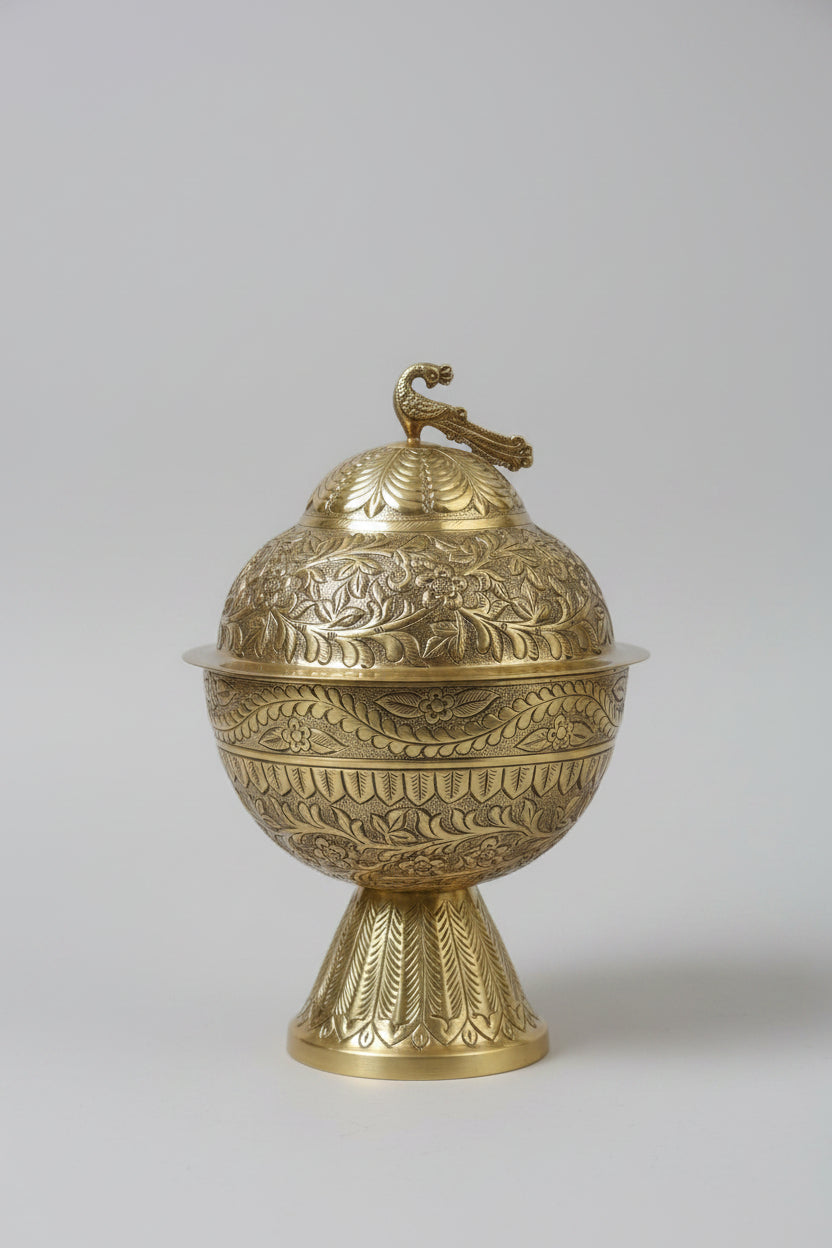 An ornate brass decorative container with a matching lid, featuring intricate floral engravings and a small bird figure on top, displayed on a dark marble surface.