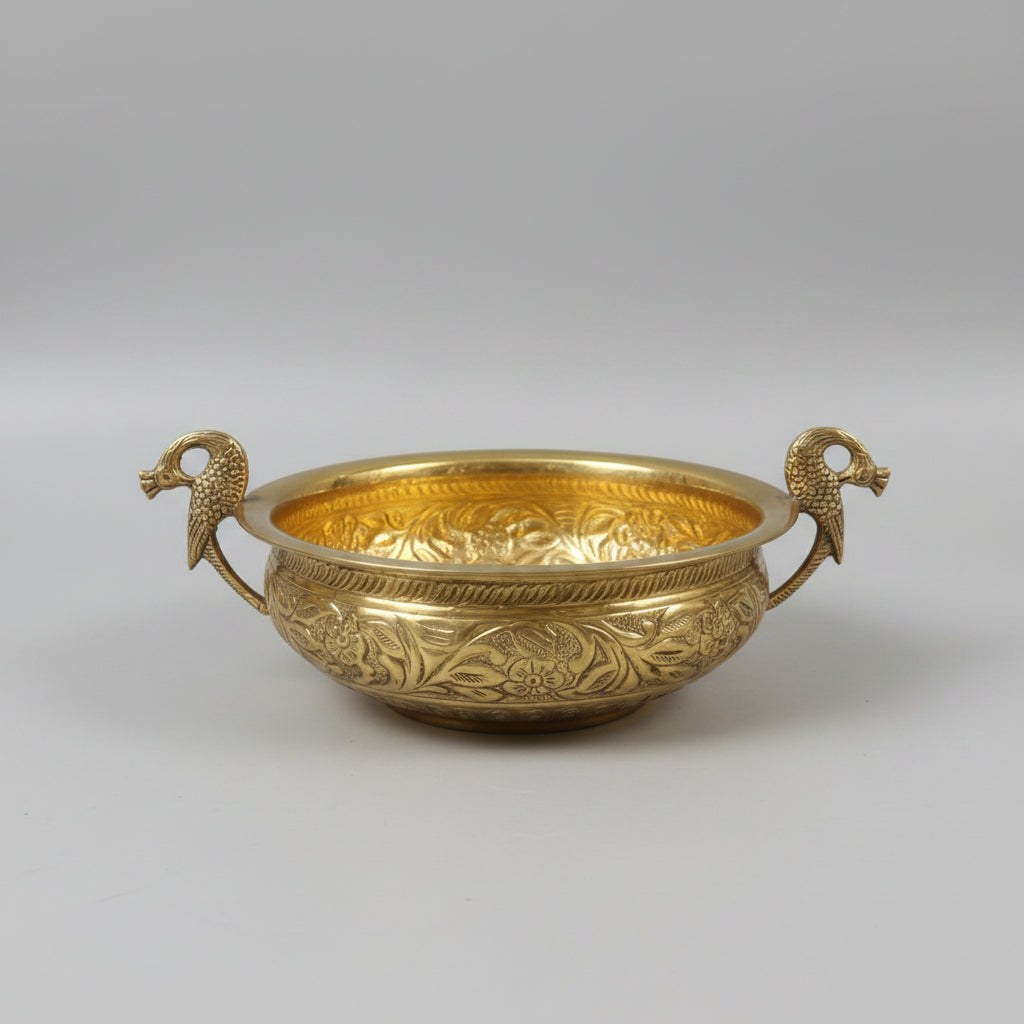 An ornate brass bowl with intricate floral engravings and two decorative bird handles, displayed on a glass table in a shop setting.