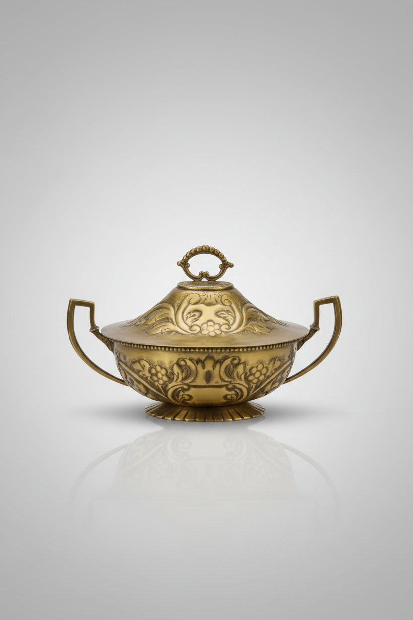 An ornate brass tureen with a lid, featuring embossed floral patterns and side handles, displayed on a glass-topped table inside what appears to be a shop.
