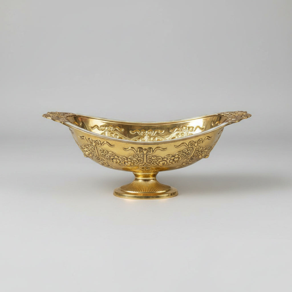 An ornate gold-colored metal serving bowl with intricate embossed floral patterns, displayed on a reflective glass tabletop in what appears to be a shop setting.