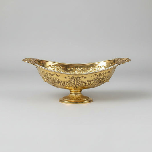 An ornate gold-colored metal serving bowl with intricate embossed floral patterns, displayed on a reflective glass tabletop in what appears to be a shop setting.