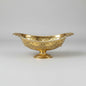 An ornate gold-colored metal serving bowl with intricate embossed floral patterns, displayed on a reflective glass tabletop in what appears to be a shop setting.