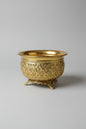 An ornate gold-colored metal incense burner with intricate floral relief patterns and three feet, displayed on a dark marble surface.