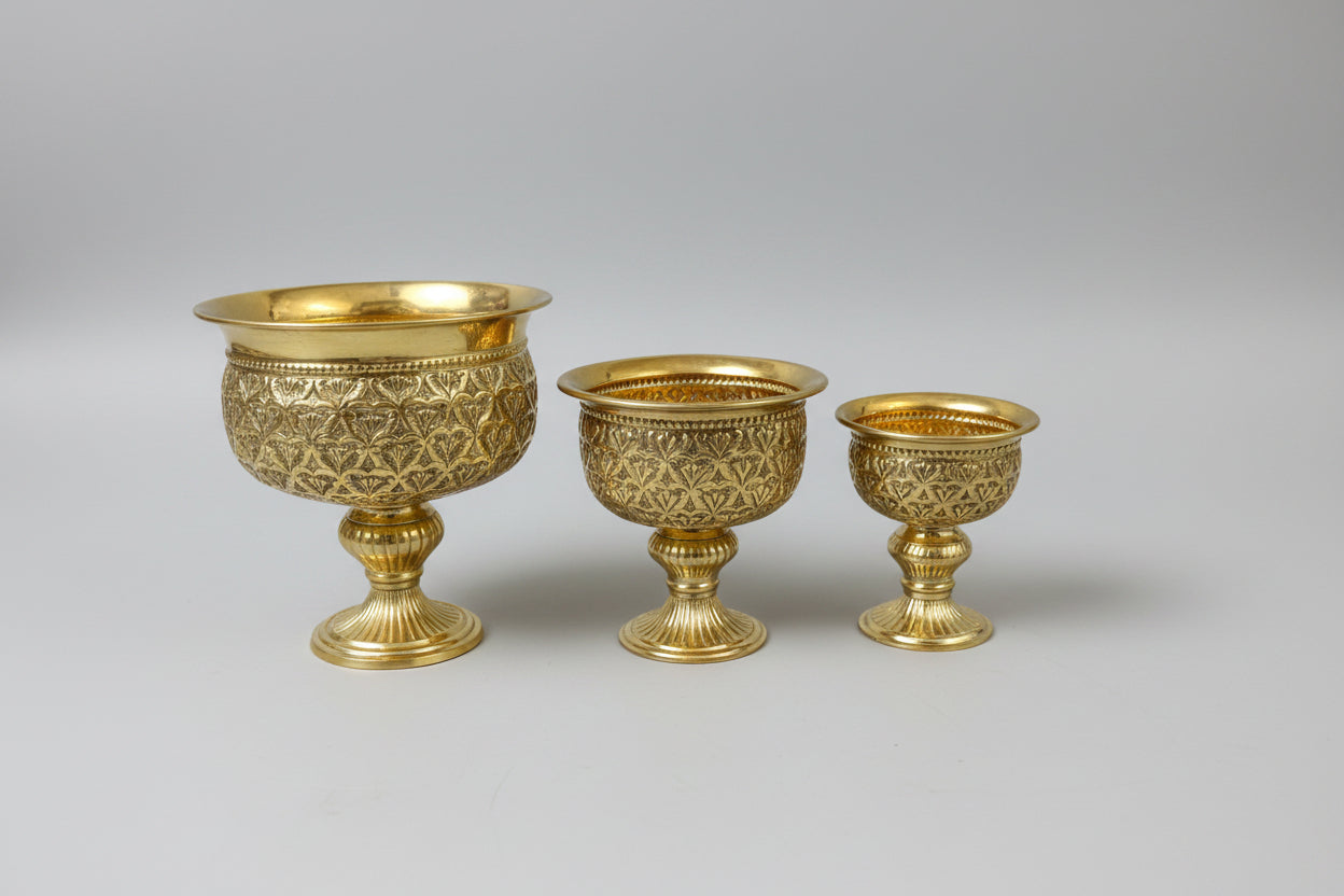 Three ornate gold-colored metal pedestal vessels of different sizes with intricate embossed patterns, displayed on a reflective dark marble table.