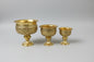 Three ornate gold-colored metal pedestal vessels of different sizes with intricate embossed patterns, displayed on a reflective dark marble table.