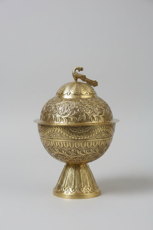 An ornate brass decorative container with a matching lid, featuring intricate floral engravings and a small bird figure on top, displayed on a dark marble surface.