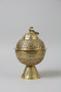 An ornate brass decorative container with a matching lid, featuring intricate floral engravings and a small bird figure on top, displayed on a dark marble surface.