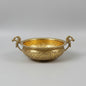 An ornate brass bowl with intricate floral engravings and two decorative bird handles, displayed on a glass table in a shop setting.