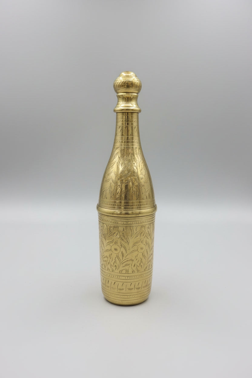 Brass bottle with intricate design on a reflective surface, with shelves of decorative items in the background.