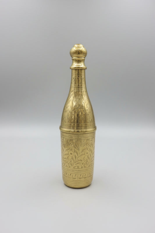 Brass bottle with intricate design on a reflective surface, with shelves of decorative items in the background.