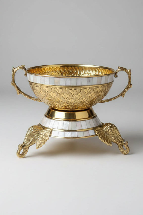 An ornate gold-toned metal bowl with mother-of-pearl inlay, featuring decorative handles and a pedestal base with elephant feet, displayed on a reflective surface in what appears to be a showroom.