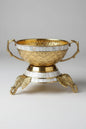 An ornate gold-toned metal bowl with mother-of-pearl inlay, featuring decorative handles and a pedestal base with elephant feet, displayed on a reflective surface in what appears to be a showroom.