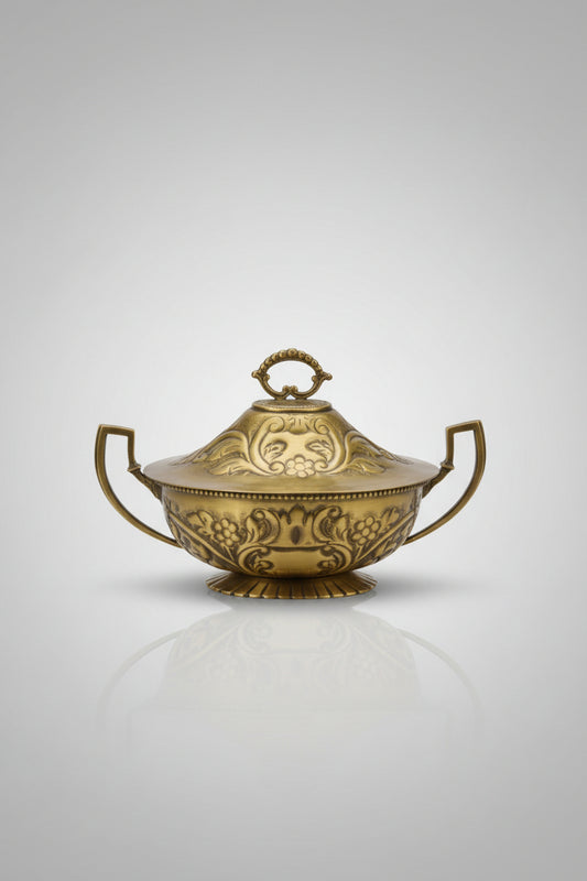 An ornate brass tureen with a lid, featuring embossed floral patterns and side handles, displayed on a glass-topped table inside what appears to be a shop.